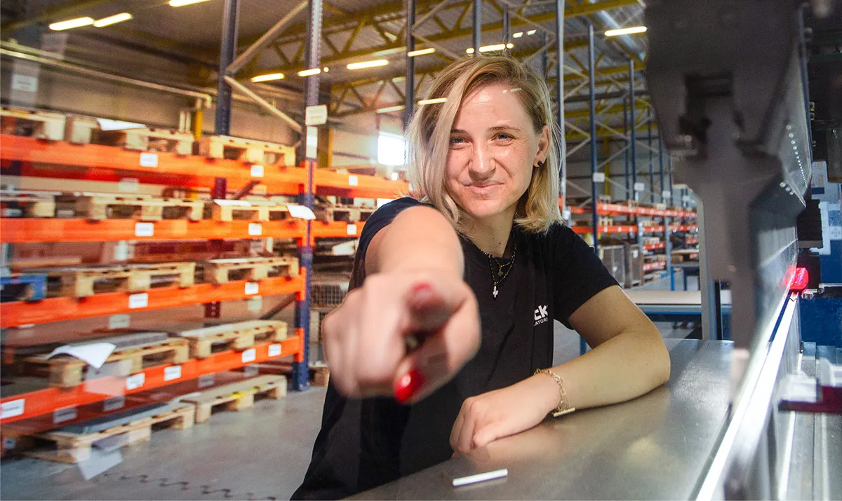 Mitarbeiterin von ruck im Logistikbereich zeigt selbstbewusst auf die Kamera, im Hintergrund Lagerregale und Produktionsumgebung.